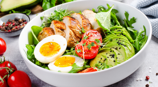 Bol de ensalada con rúcula, pollo a la plancha, aguacate, tomate cherry y huevo cocido, decorado con semillas de sésamo.
